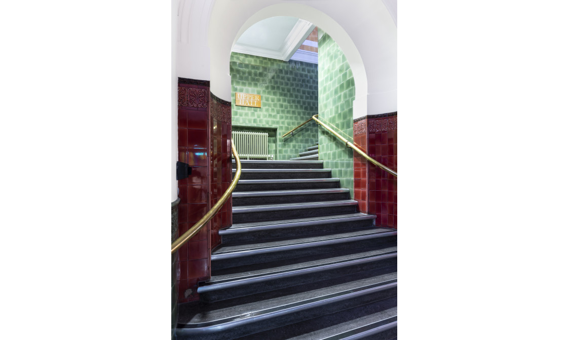 A staircase at Bishopsgate Institute. The stairs are black with gold bannisters. There are red tiles on the wall leading up to another staircase with green tiles.