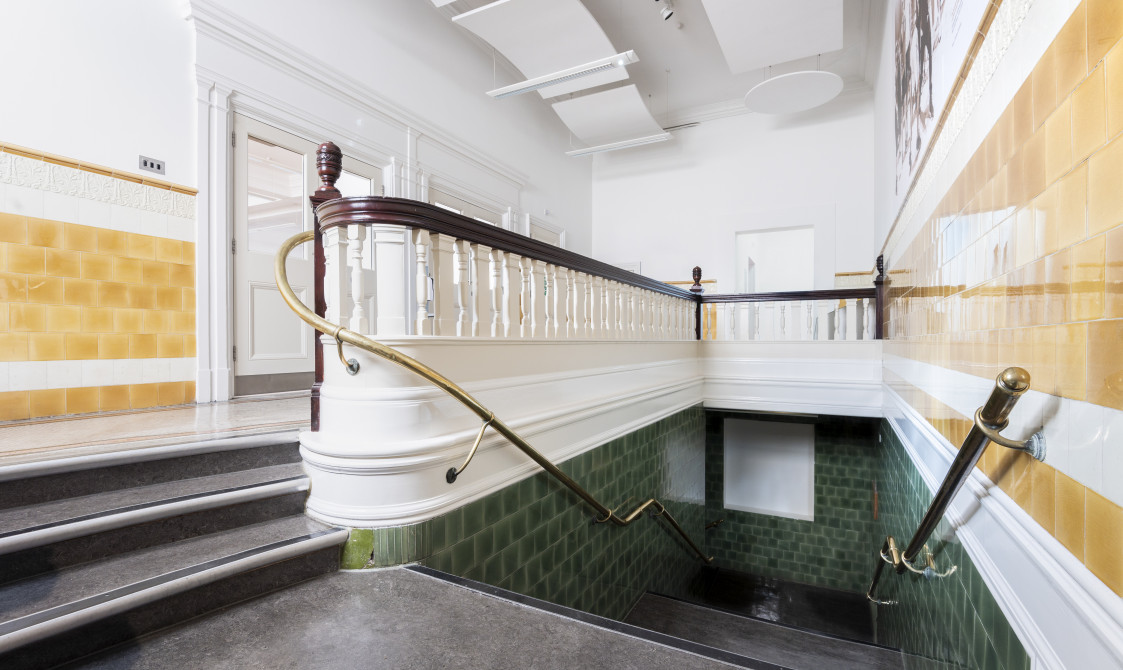 The landing to the Upper Hall at Bishopsgate Institute. There are yellow tiles on the walls and green tiles along the staircase. There are also wooden bannisters on either side of the staircase, which leads up to a landing.