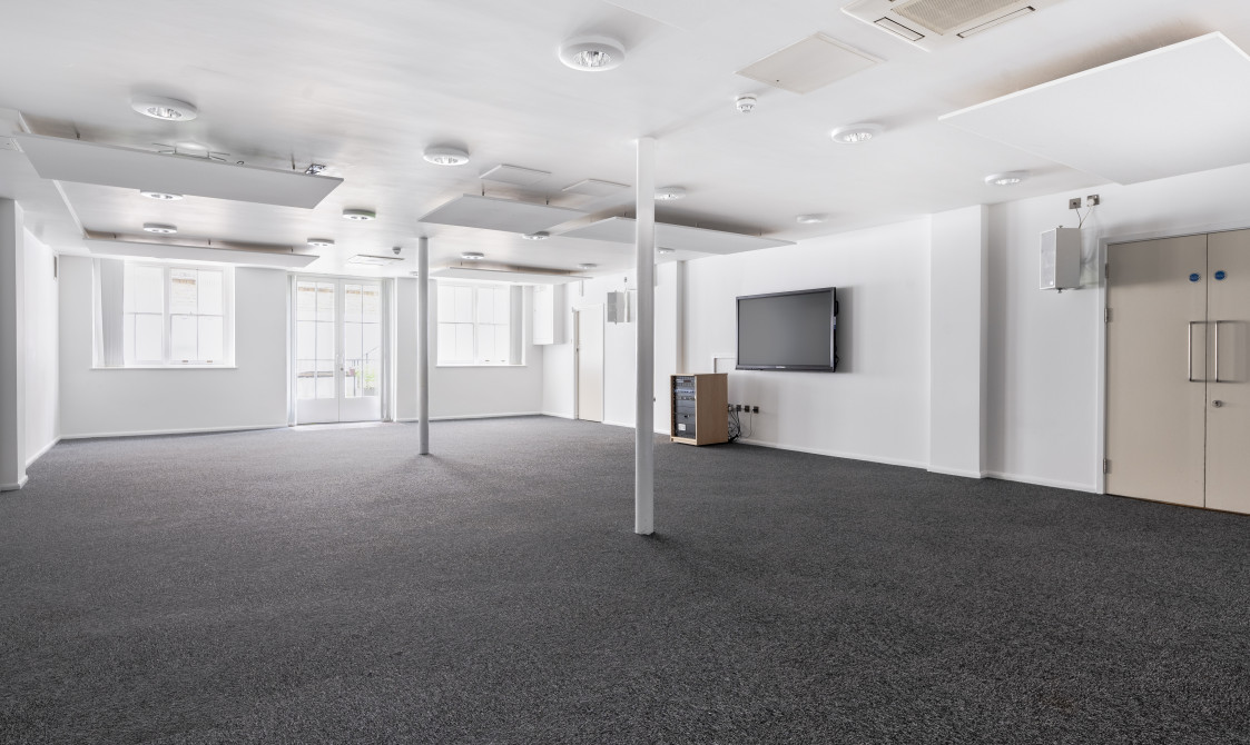 A photograph of Bishopsgate Institute's Courtyard Room in central London. The floor is carpeted and the walls are white, with a screen mounted to the walls.