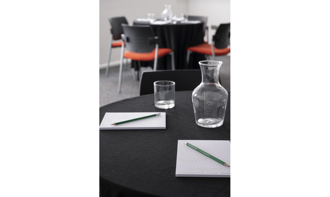 A photograph of Bishopsgate Institute's Courtyard Room in central London. The image is focused on a table top with pens and paper on it and a glass of water.