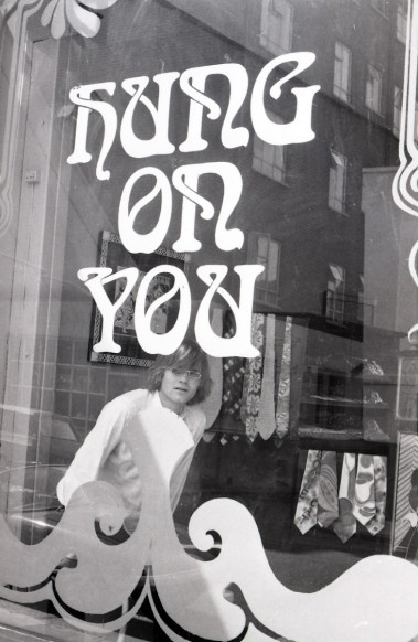 A black and white photo from the 1960s of a man stood inside a shop looking out the window. Stickers on the glass of the window say 'Hung on You'