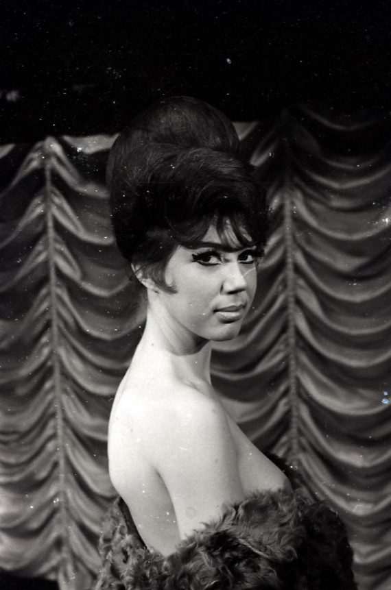 A black and white photograph of a stripper in the 1960s. She is naked, but draped in a fur. She has a large beehive hairstyle and is stood in front of a theatrical curtain.