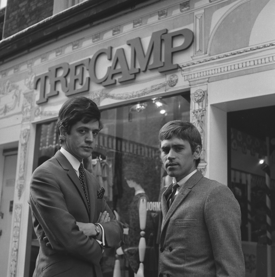 Black and white photo from the 1960s of two young men dressed in suits, with similar mod haircuts, looking stern. They are stood outside a clothing shop called Tre Camp.
