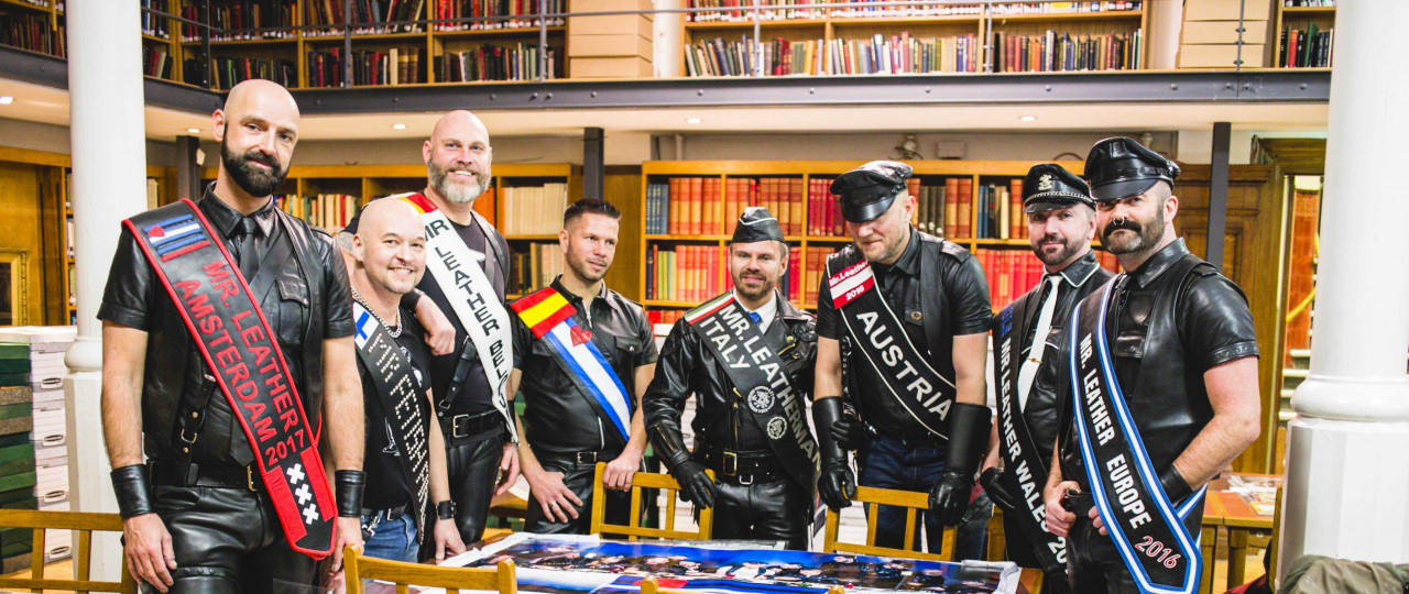 A photo from the opening of the UK Leather & Fetish Archive at Bishopsgate Institute. Eight men are standing round a table in the Library, dressed up in leather.