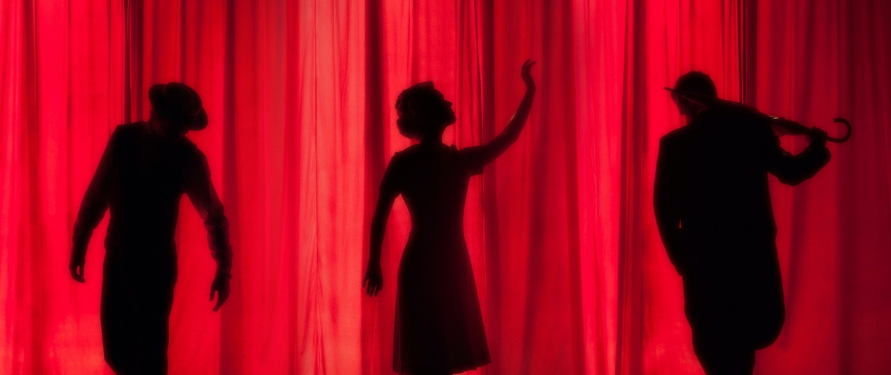 Image of the silhouettes of 3 performers in front of a low lit red curtain backdrop.