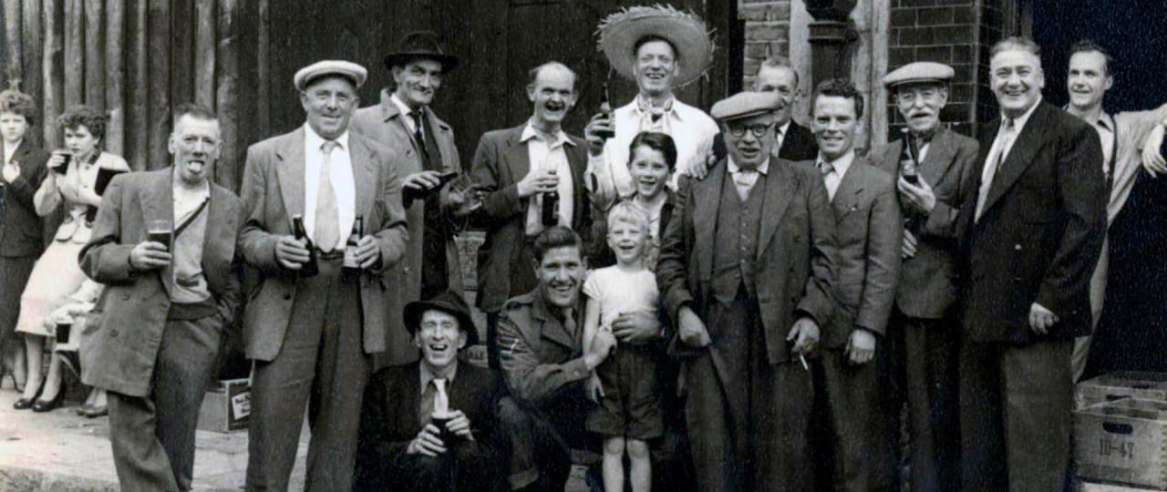 A black and white photo of people living in the East End, who are holding drinks and smiling at the camera. Most of the people in the photo are men in suits and flat caps, with a few children also in the photo.