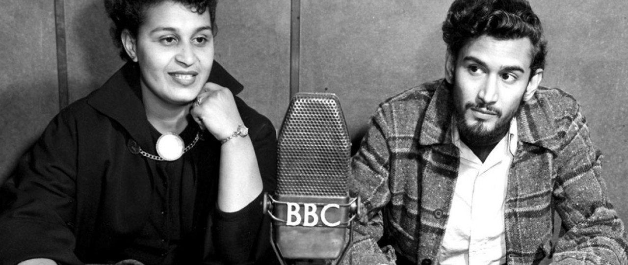 Black and white photo of Samuel Selvon and Pauline Henriques in front of a BBC mic.