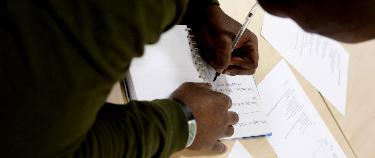 A landscape photo of a student making notes in a class. He is writing in a notebook, with the pen in his left hand. The photo is taken over his shoulder.