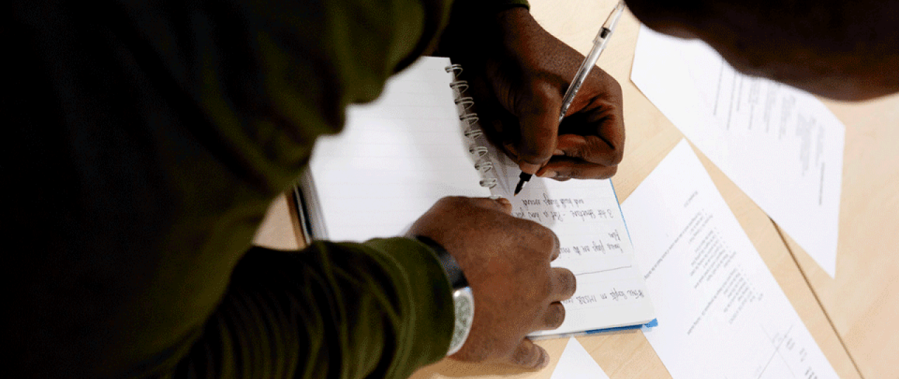 A student is making notes on a pad of paper at a creative writing session. The photo is taken over their shoulder. Their pen is in their left hand side.