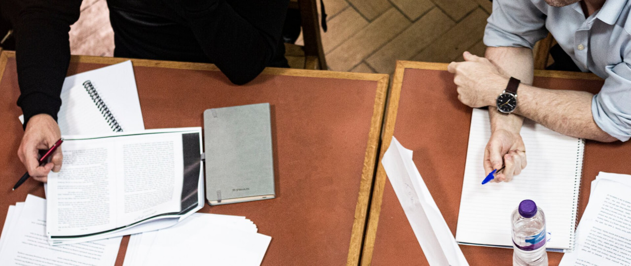 Two people are writing in notebooks on a table. They also have photocopies of book extracts in front of them. The photo is taken from above.