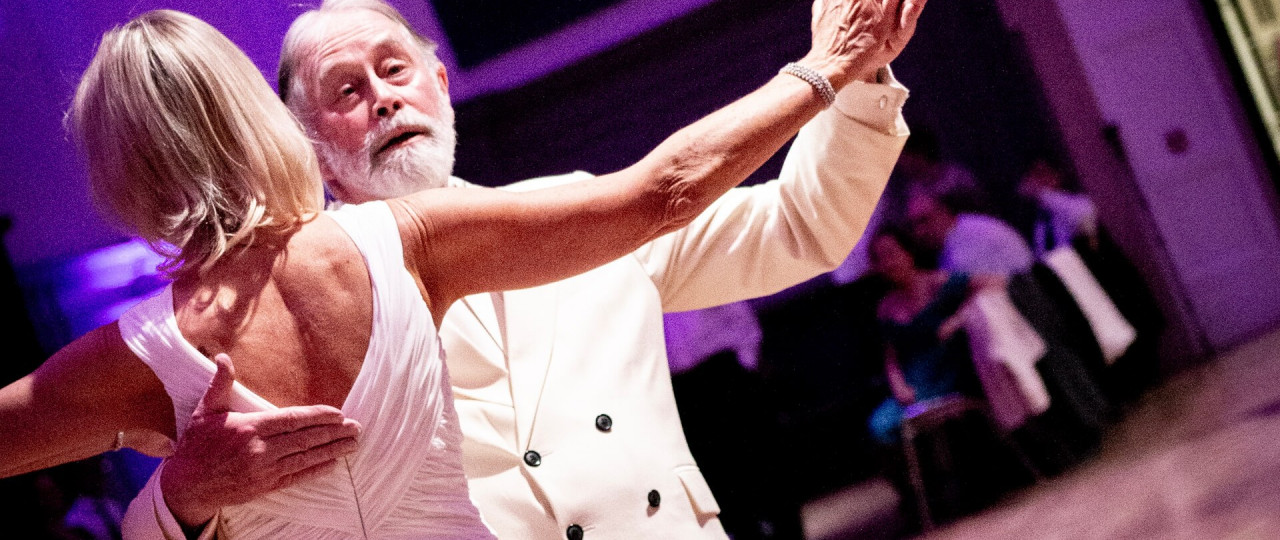 A picture of a man and a woman slow dancing. They are in a Foxtrot hold and the photo is taking from behind the woman as they dance. They are in the Great Hall, which has pink lighting.