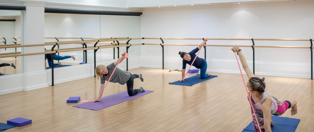 A Pilates class at Bishopsgate Institute. Students and a tutor in a large studio with ballet bars in the background. Students and a tutor are balancing on one hand, one knee whilst stretching a band on top of a yoga mat.