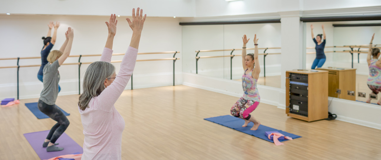 A Pilates class at Bishopsgate Institute. Students and a tutor are standing with knees slightly bent with hands above their heads whilst on a yoga mat. There are also yoga bands and cushions in the room.