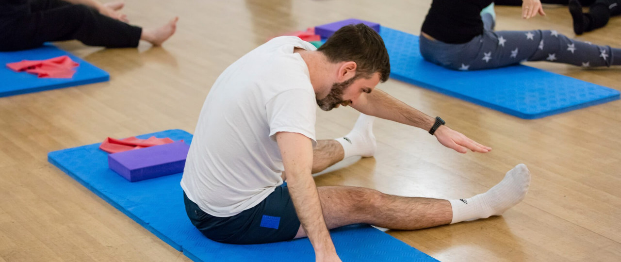 A pilates class at Bishopsgate Institute. Students are sitting on blue mats, and are in a pose where they are reaching over to touch their toes with their hands. Cushions, straps, and balls are also being used in the class.