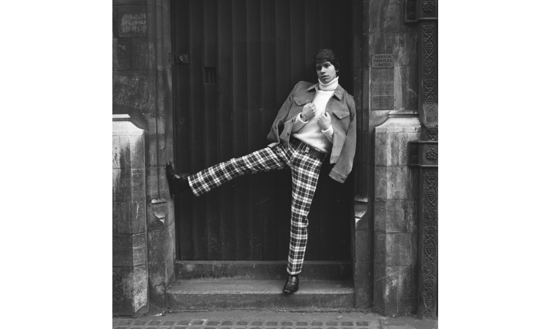 A man is standing in a doorway, wearing chequered trousers, a turtleneck top and a jacket. He has his right leg kicked up in the air. The image is black and white and was taken in the 1960s.