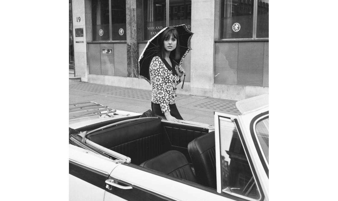 A woman is standing in front of a car, wearing a top with a circular pattern, a black skirt, and is holding an umbrella. The image is in black and white and taken during the 1960s.