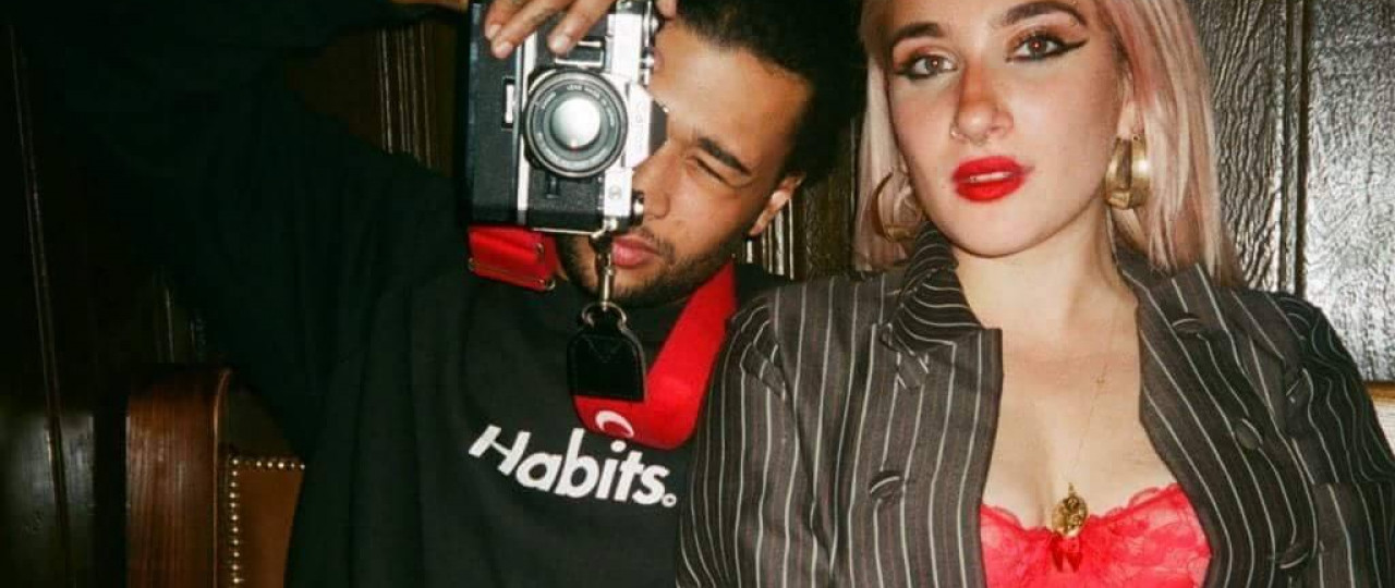 The two people who run London Vagabond. The man on the left is taking a photo, and the woman is sitting next to him, both looking at the camera.