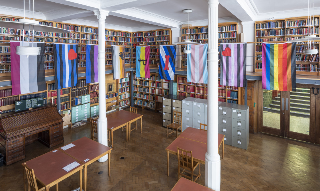 A photograph of Bishopsgate Institute's Researchers' Room. There are flags hanging at the top of the room, including LGBTQ+ flags. There are bookshelves around the room, and wooden tables and chairs in the centre of the room.