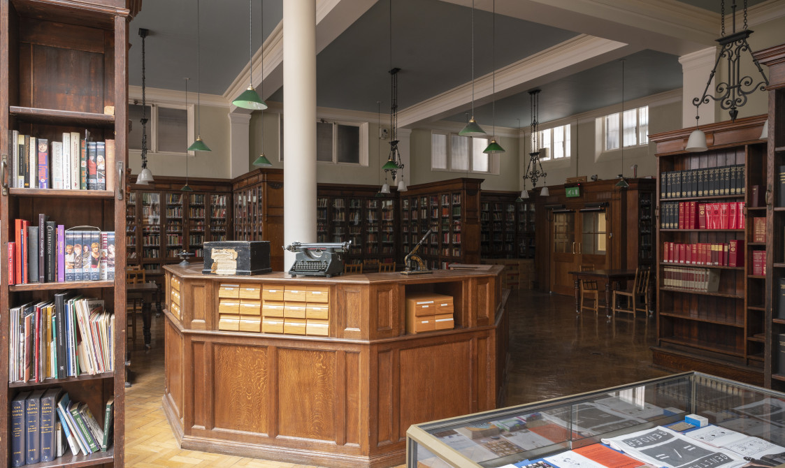 A photograph of Bishopsgate Institute's Library in central London. There is a desk in the centre of the room and bookshelves around the edges of the room. There is also a glass cabinet displaying materials near the desk.