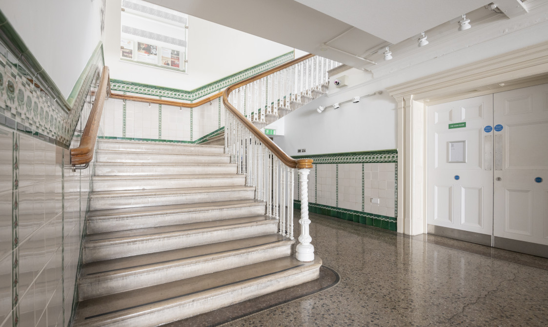 A photograph of the basement staircase at Bishopsgate Institute in central London. The staircase is white with a wooden banister. Green tiles go up along the staircase.