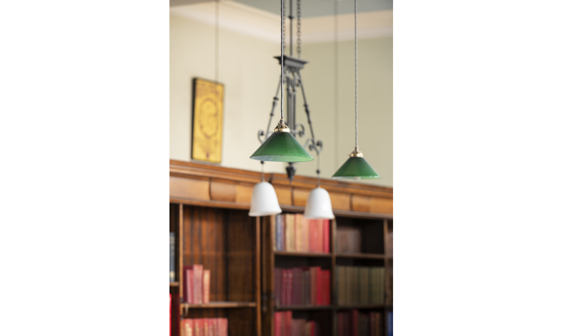 A photography of two hanging lights in Bishopsgate Institute's Library in central London. The lampshades are green and bookshelves are behind the lights.