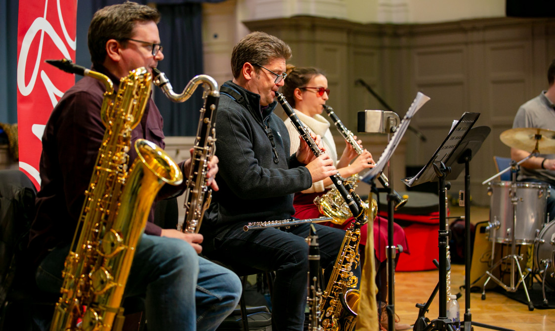 Two musicians are playing the saxophone, and two others are playing wind instruments, with a drummer in the background.