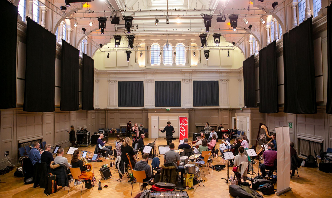 An orchestra rehearsal in Bishopsgate Institute's Great Hall in central London. A conductor is at the back of the room, with musicians around them. A LMTO banner is at the back of the room.