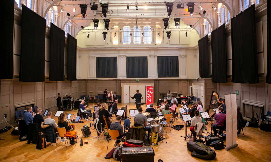 An orchestra rehearsal in Bishopsgate Institute's Great Hall in central London. A conductor is at the back of the room, with musicians around them. A LMTO banner is at the back of the room.