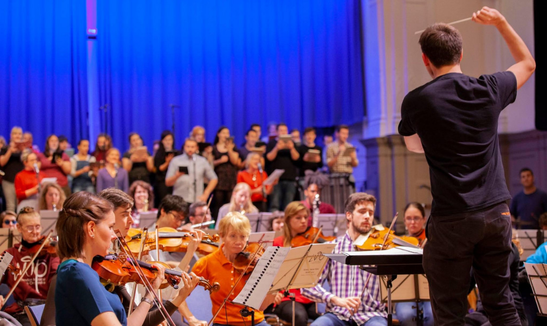 An orchestra rehearsal in Bishopsgate Institute's Great Hall. A conductor is in the right of the photo with musicians in front of him, playing villains and other instruments. There are singers on stage.