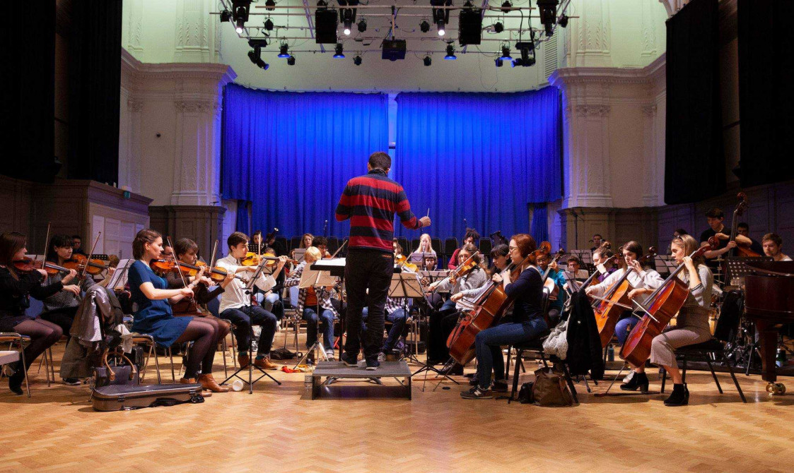 Orchestra rehearsals in Bishopsgate Institute's Great Hall. A conductor is standing in the middle with musicians around them in a semi circle. They are are playing the cello, violin and other instruments. A stage is behind them with a blue curtain.